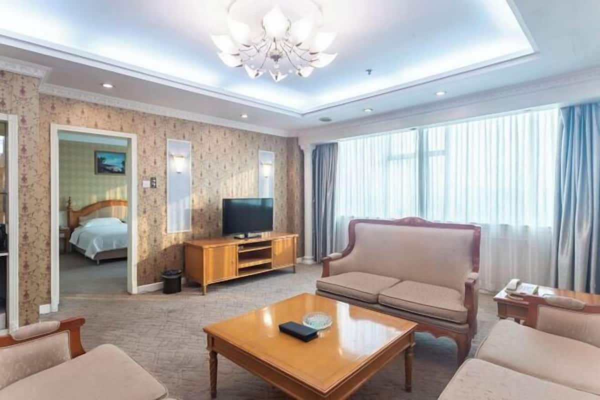 A hotel room with a sofa, a television on a wooden stand, a coffee table, and a large window with curtains.