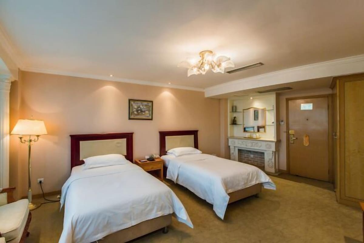 A hotel room with two beds, a bedside table, a lamp, a mirror, and a door.