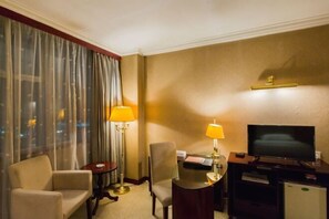 A hotel room with a bed, a desk with a lamp, a television, and a chair.