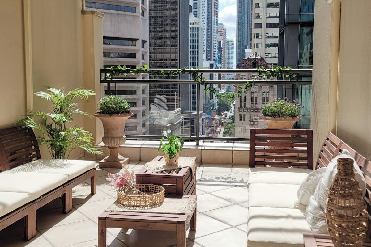 Un balcón con vista a edificios altos, amoblado con sillas y mesas de madera, y decorado con plantas en macetas.