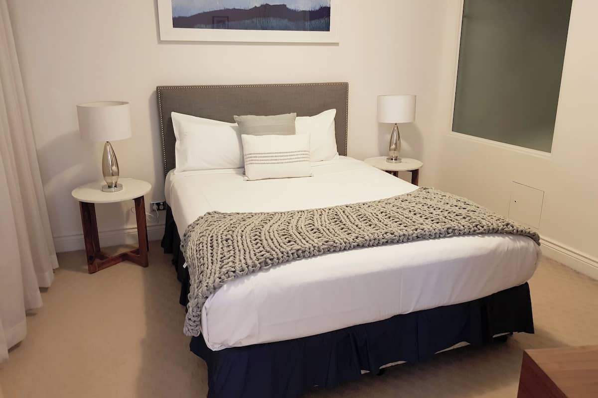 Un dormitorio ordenado con una cama grande, dos mesitas de noche, un cuadro en la pared y una ventana.