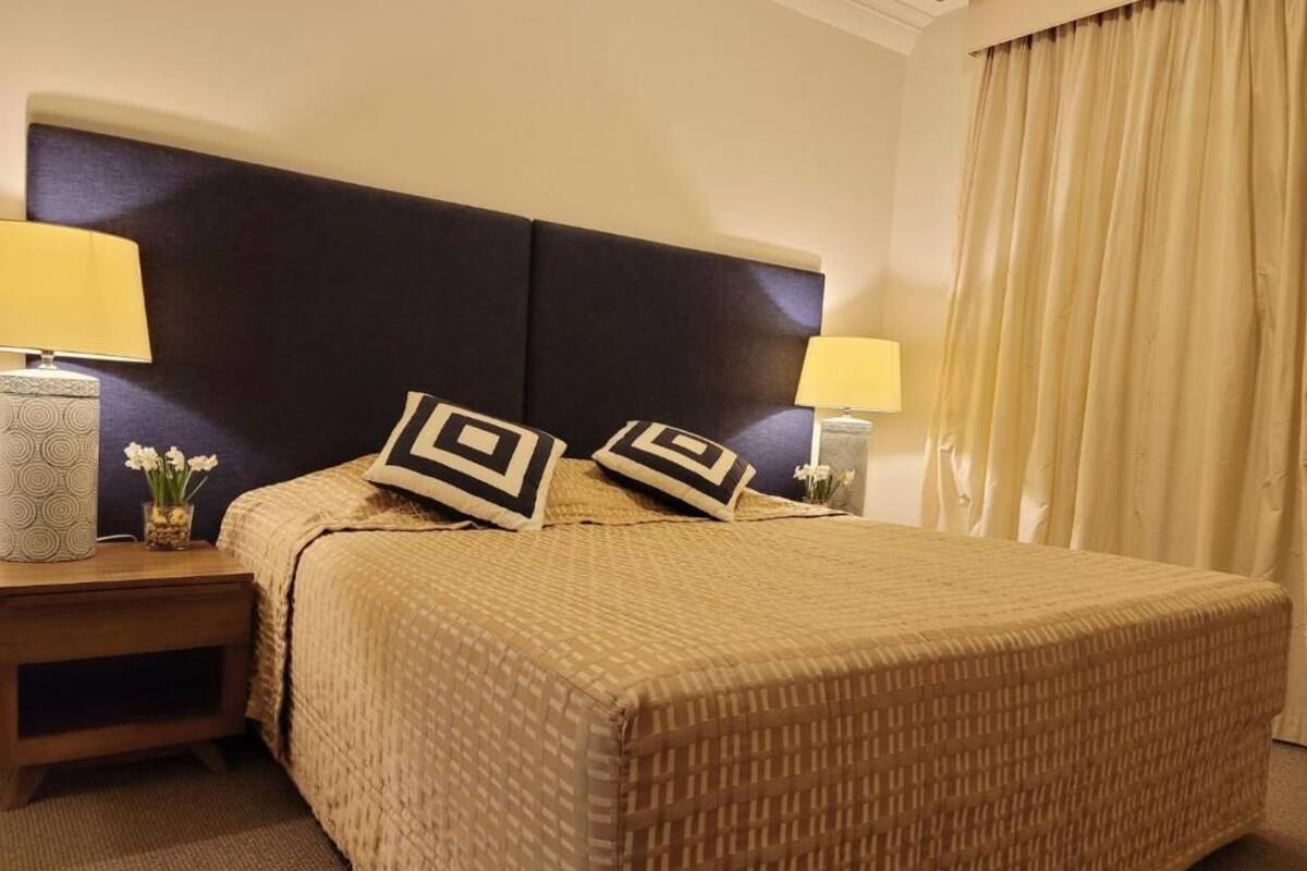 Una habitación de hotel con una cama grande, dos mesitas de noche con lámparas, un cabecero y una cortina.