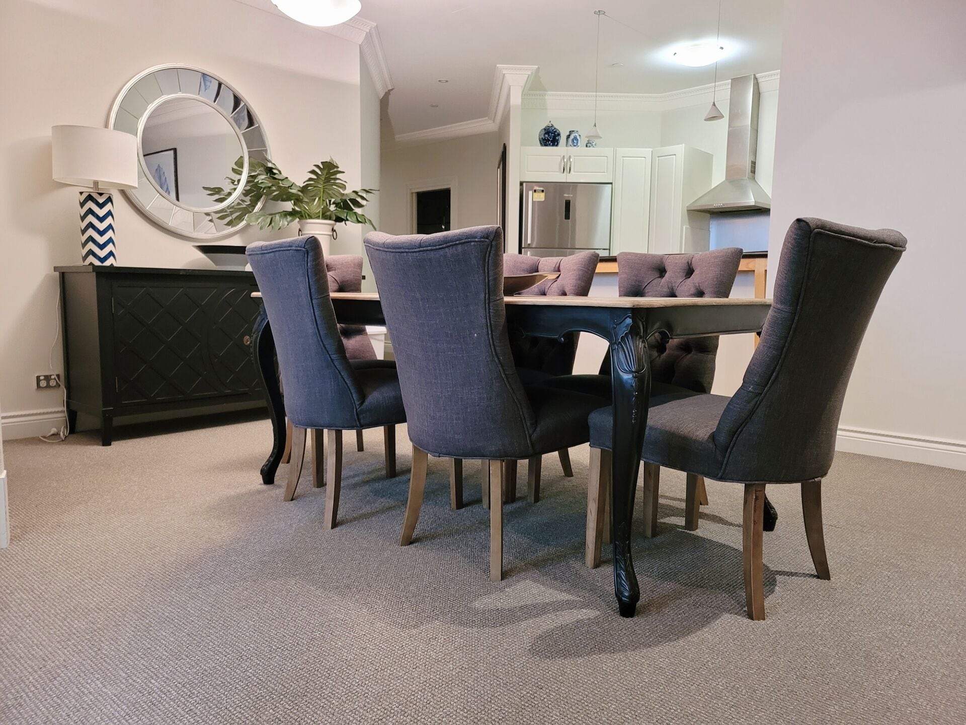 Un coin repas comprenant une table ronde, quatre chaises, un meuble noir, un miroir et une plante en pot.