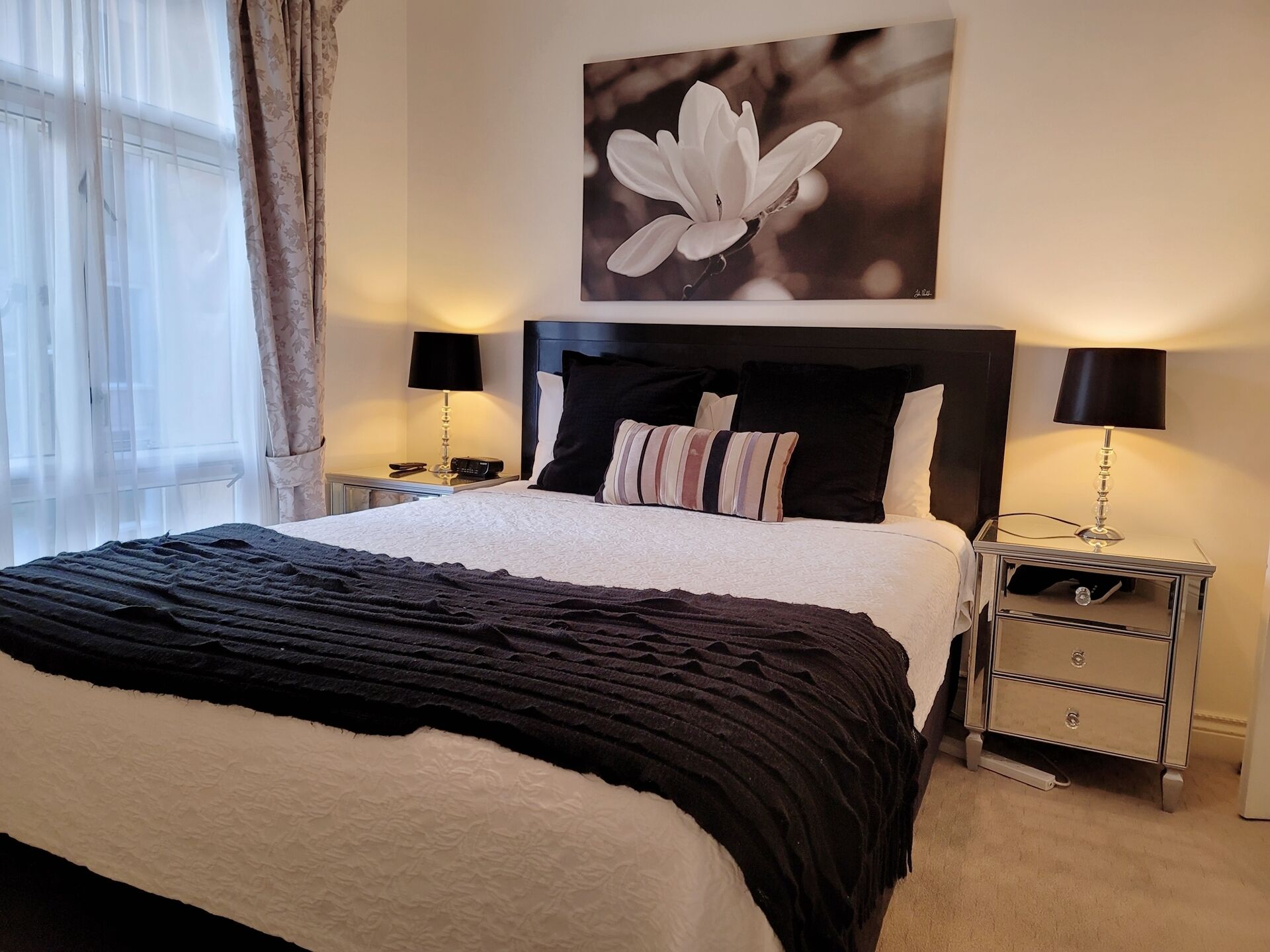 Un lit bien fait, avec un oreiller à rayures noires et blanches, un plaid noir et un tableau représentant une fleur accroché au mur. Il y a deux tables de chevet avec des lampes et un petit tiroir.
