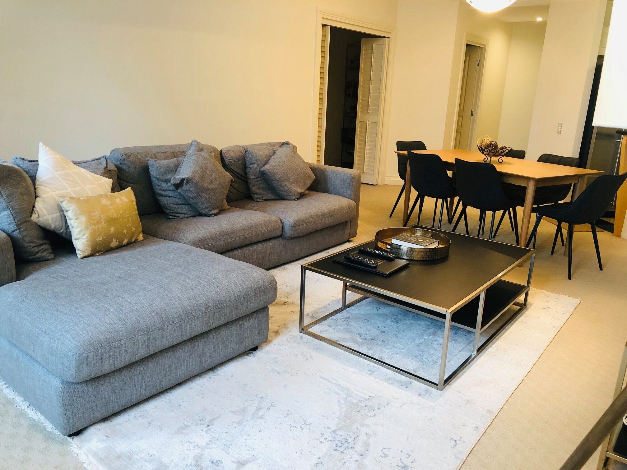 Ein modernes Wohnzimmer mit einem grauen Ecksofa, einem Couchtisch mit Glasplatte und einem Essbereich mit Holztisch und schwarzen Stühlen.