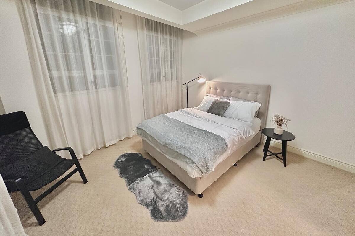 Un dormitorio con cama, una silla, una mesita y una alfombra en el suelo.
