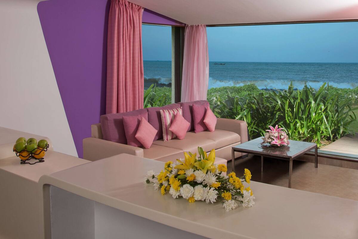 Un salon moderne avec vue sur l’océan, un canapé agrémenté de coussins roses, une table basse ornée de fleurs et une plante posée sur le rebord de la fenêtre.