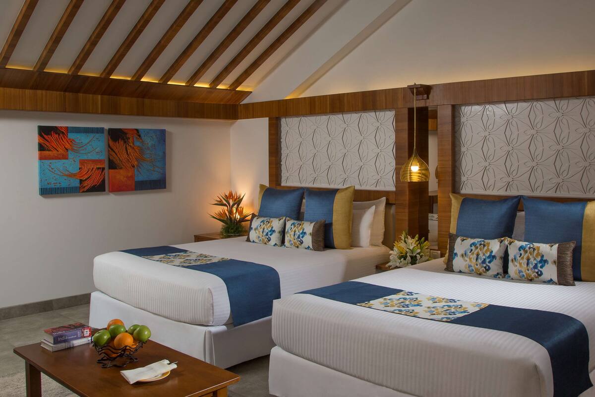 Une chambre d’hôtel avec deux lits, des poutres en bois au plafond, des panneaux décoratifs sur les murs et une petite table sur laquelle se trouve un panier de fruits.