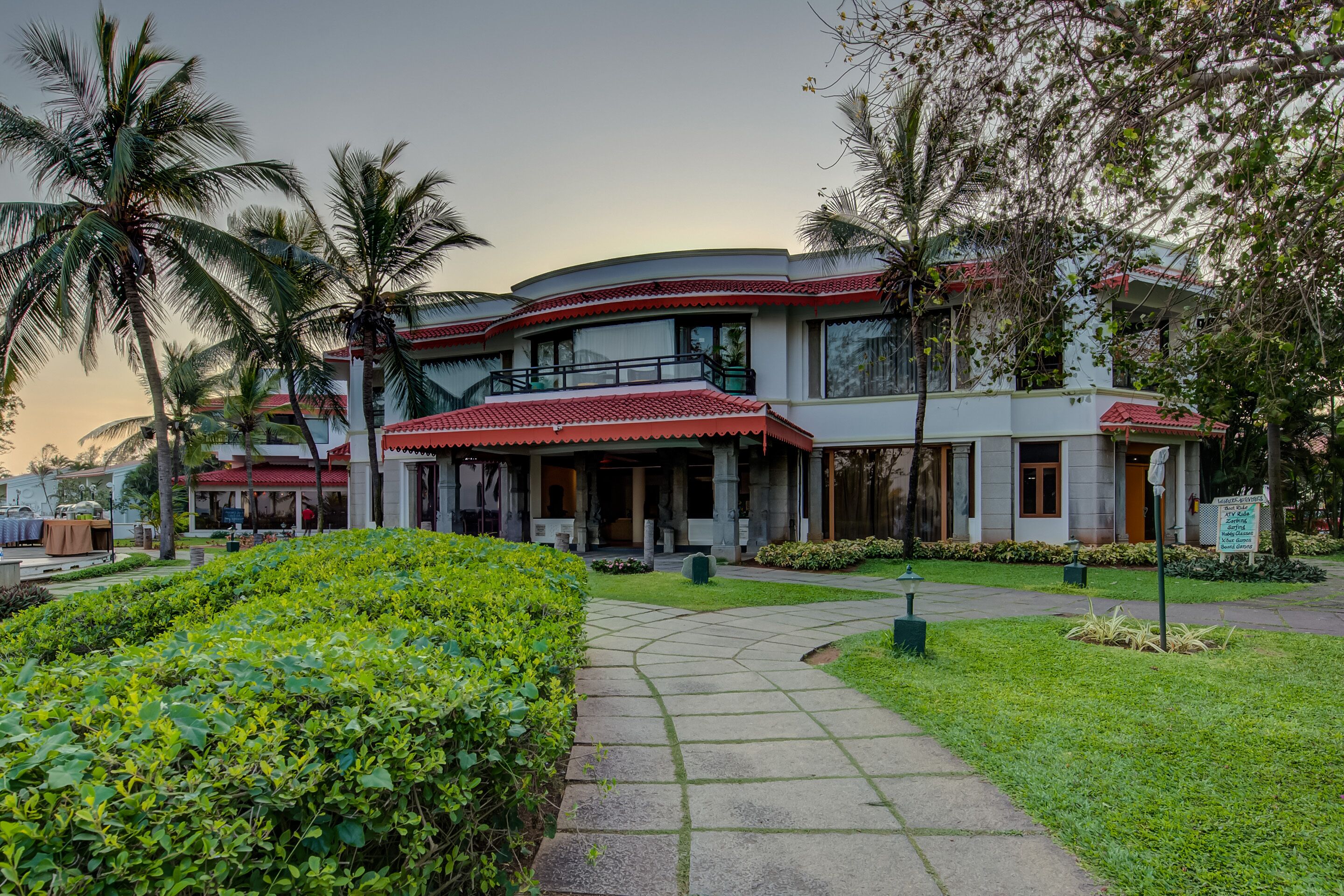 Un hotel con fachada curva, varios balcones y un jardín cuidado con palmeras y setos.