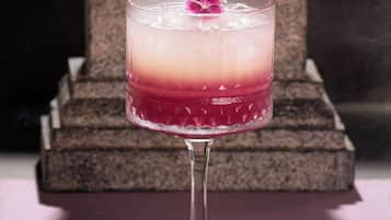 Een cocktail in een longdrinkglas met een kleurovergang, een roze bloem als garnering en een stenen structuur op de achtergrond.