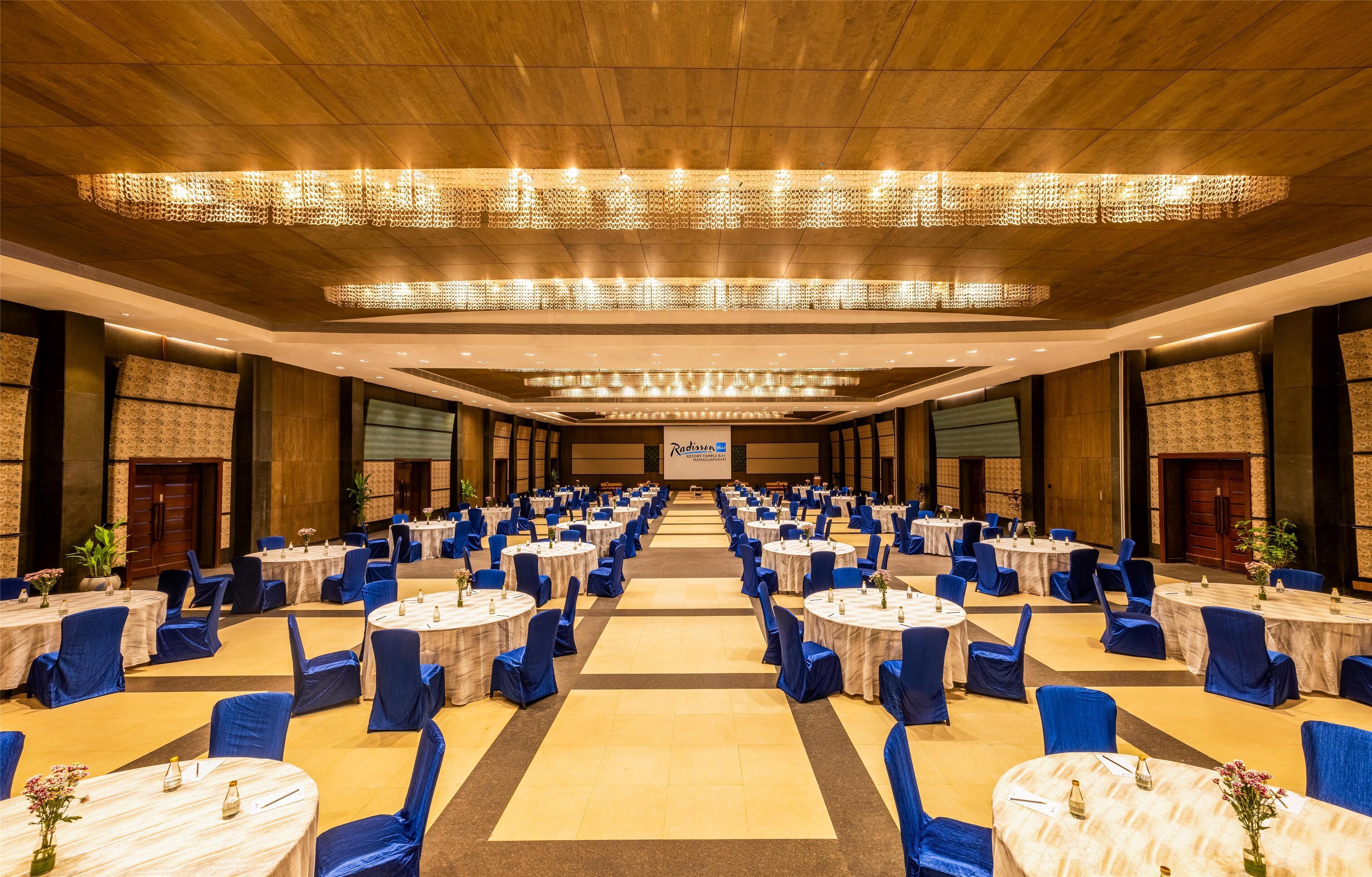 A large banquet hall with round tables and blue chairs, a wooden ceiling, and a stage at the front.