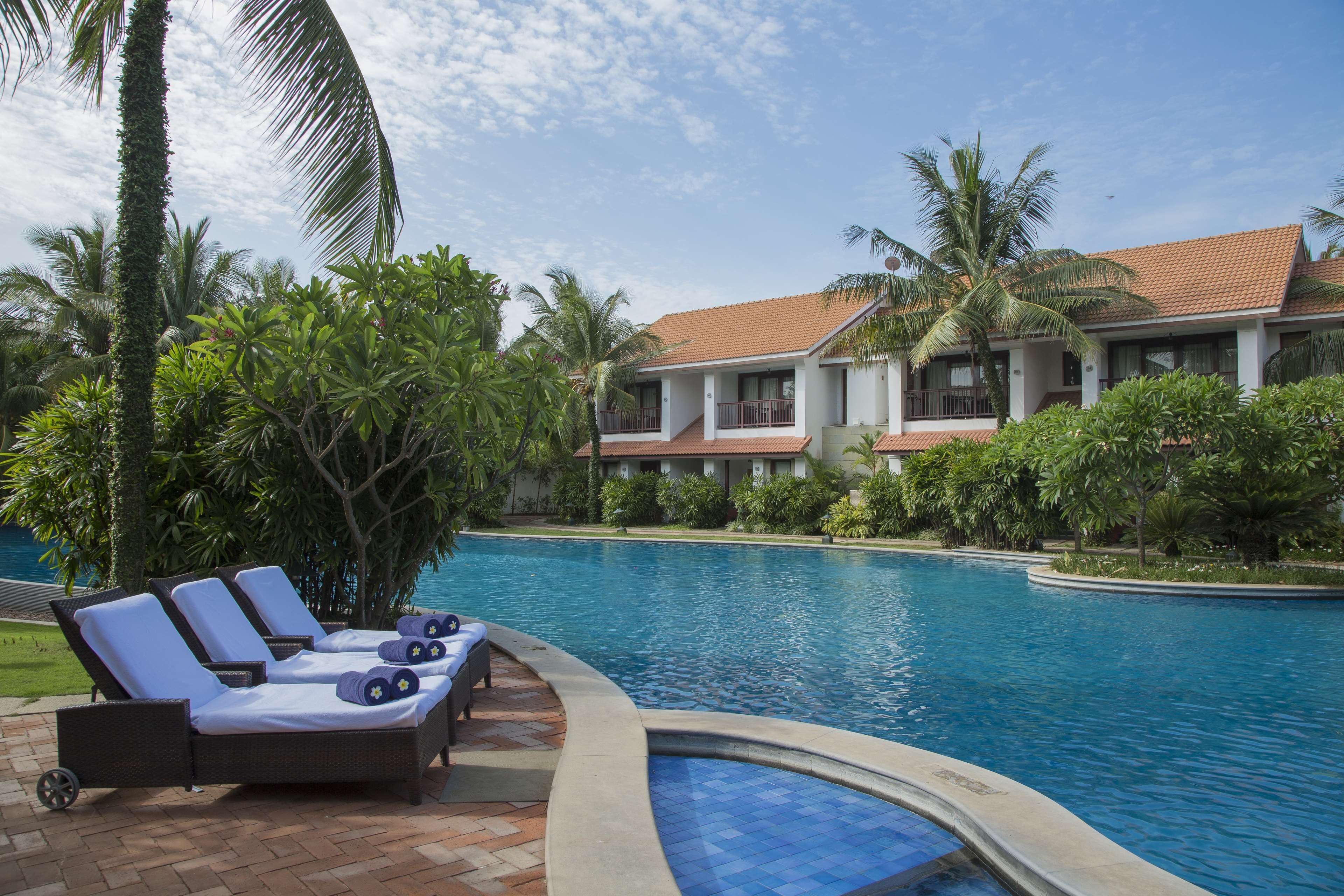 A pool area with lounge chairs, palm trees, and a building with a tiled roof.