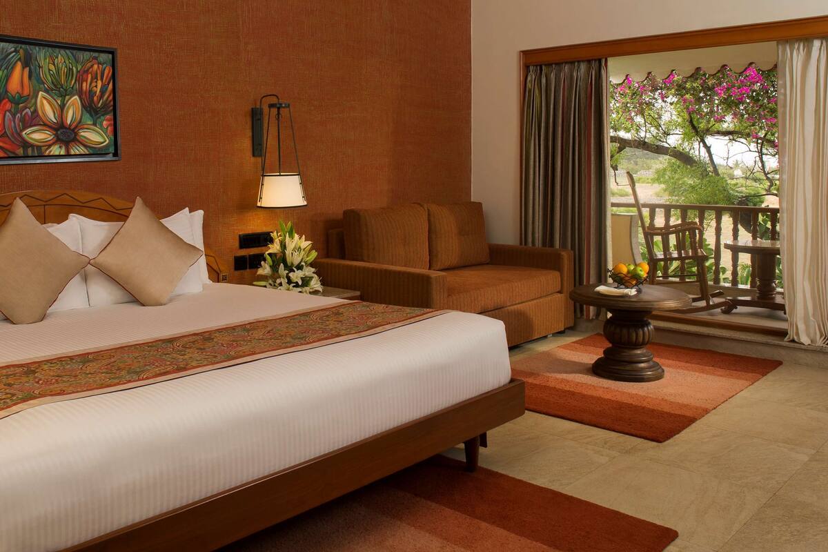 Une chambre d’hôtel avec un grand lit, un canapé, une table avec des fruits, un tableau au mur et une vue sur l’extérieur.