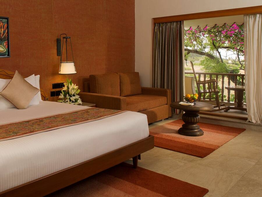 A hotel room with a large bed, a sofa, a table with fruit, a painting on the wall, and a view of the outdoors.