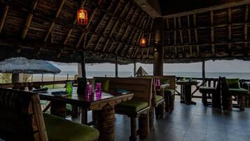 An outdoor dining area with wooden furniture and thatched roof. Tables are set with green cushions and various items.