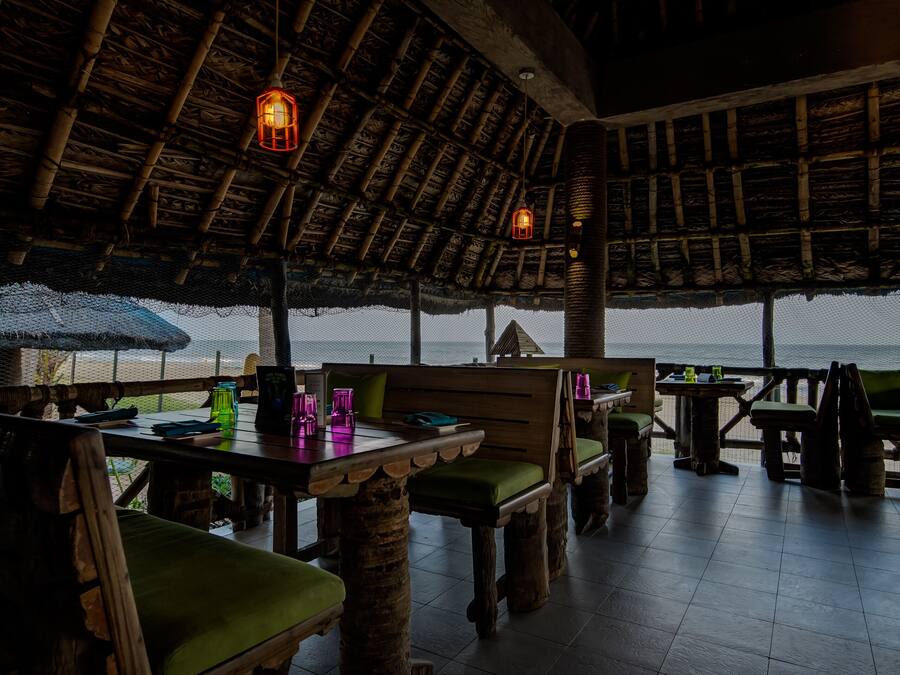 An outdoor dining area with wooden furniture and thatched roof. Tables are set with green cushions and various items.