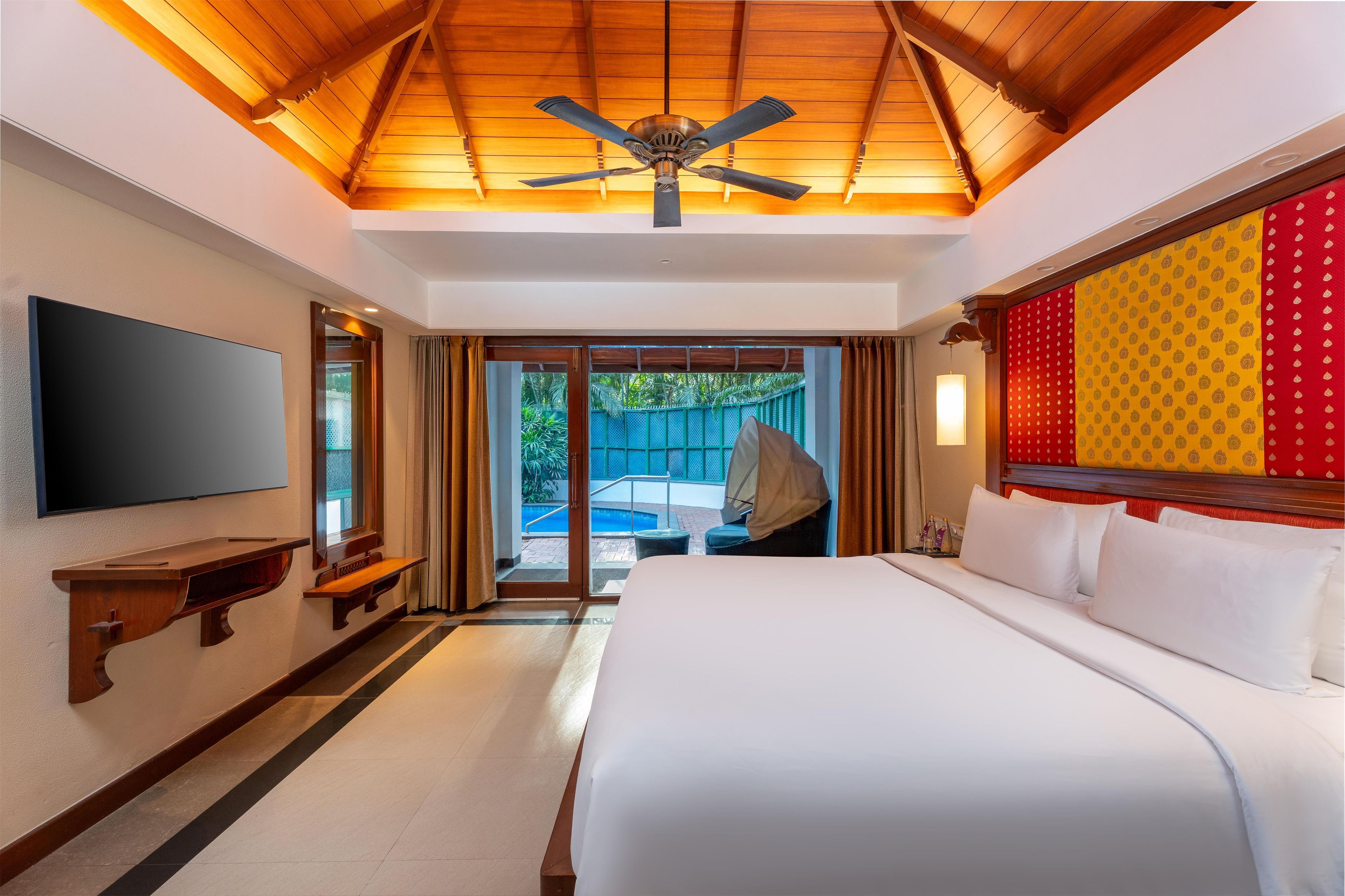 A hotel room with a bed, a flat-screen TV, a ceiling fan, and a view of a swimming pool.