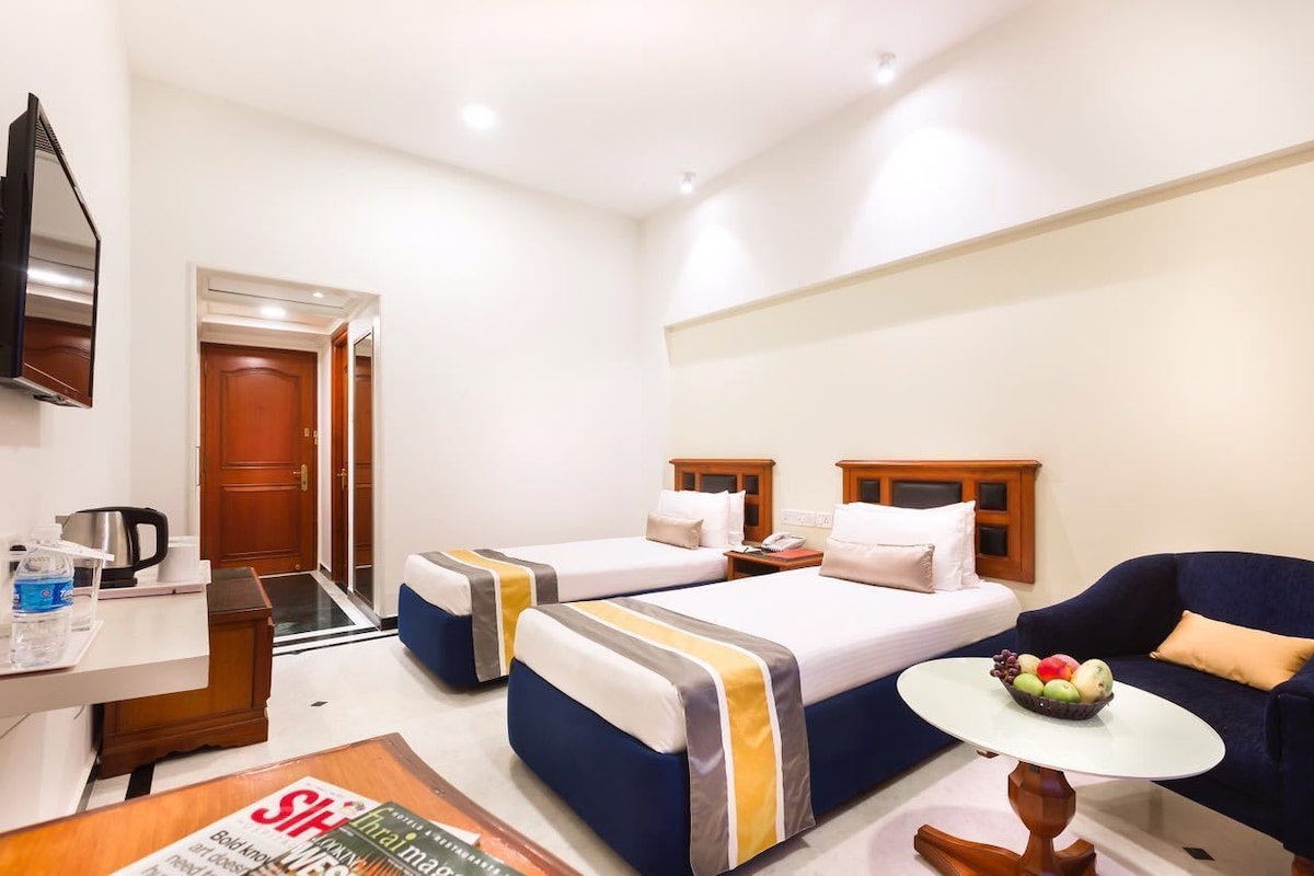 A hotel room with two beds, a sofa, a small table with fruit, a TV, and a desk with a kettle.
