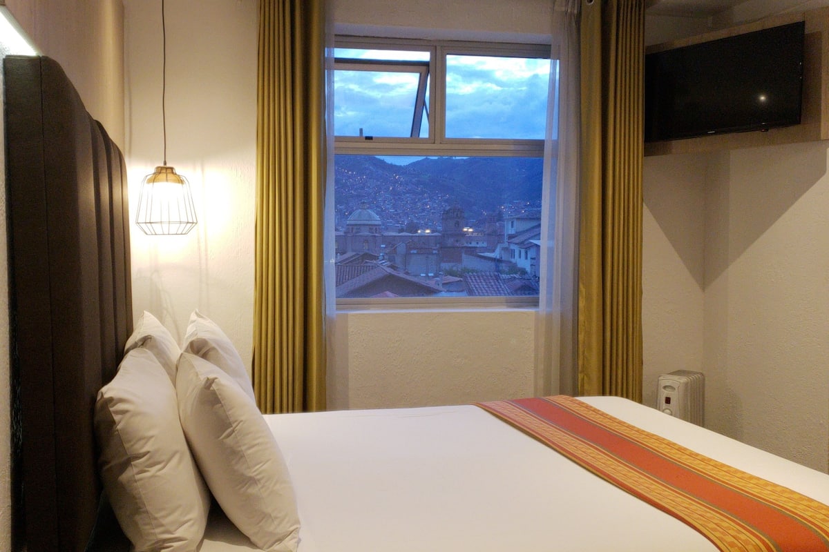 A hotel room with a bed, a window with curtains, a television mounted on the wall, and a light fixture above the bed.