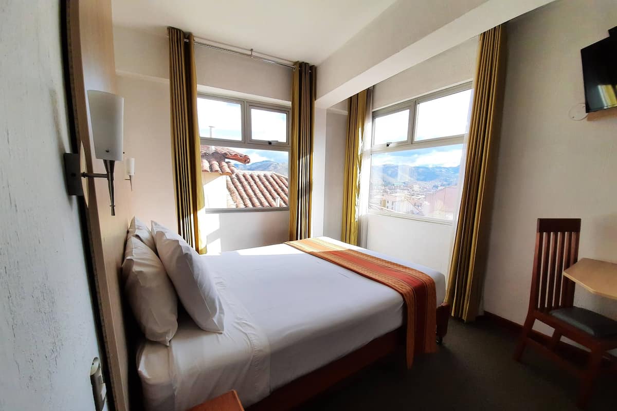 A hotel room with a bed, a desk, and a chair. There are windows with curtains, a view of the cityscape, and a wall-mounted TV.