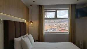 A hotel room with a large bed, a view of a cityscape through a window, and a pendant light. - Sueños del Inka Hotel (Cusco)