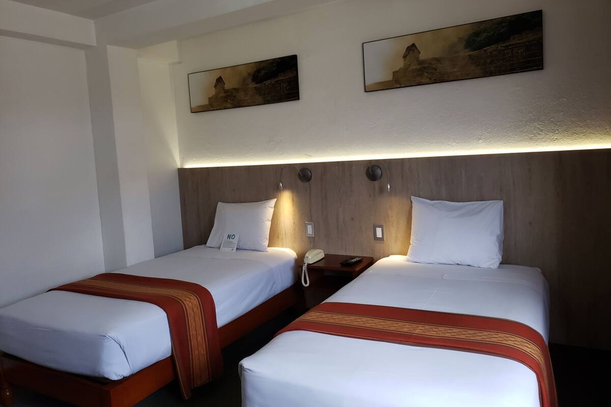 A hotel room with two single beds, each with a wooden headboard, white bedding, and a red decorative trim. There are two wall-mounted paintings above the beds, a bedside table with a telephone, and a wall-mounted light fixture.