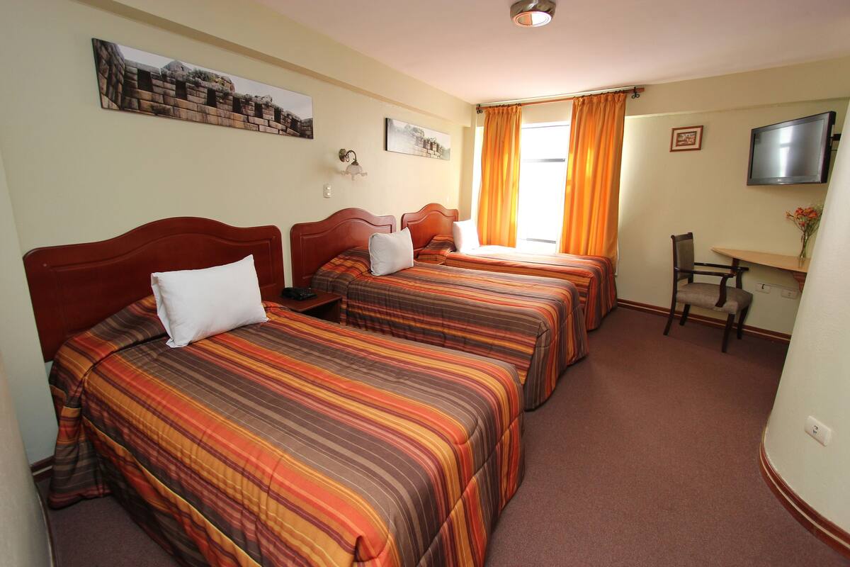 Une chambre d’hôtel avec trois lits, un bureau, une chaise, une télévision et une fenêtre avec des rideaux.