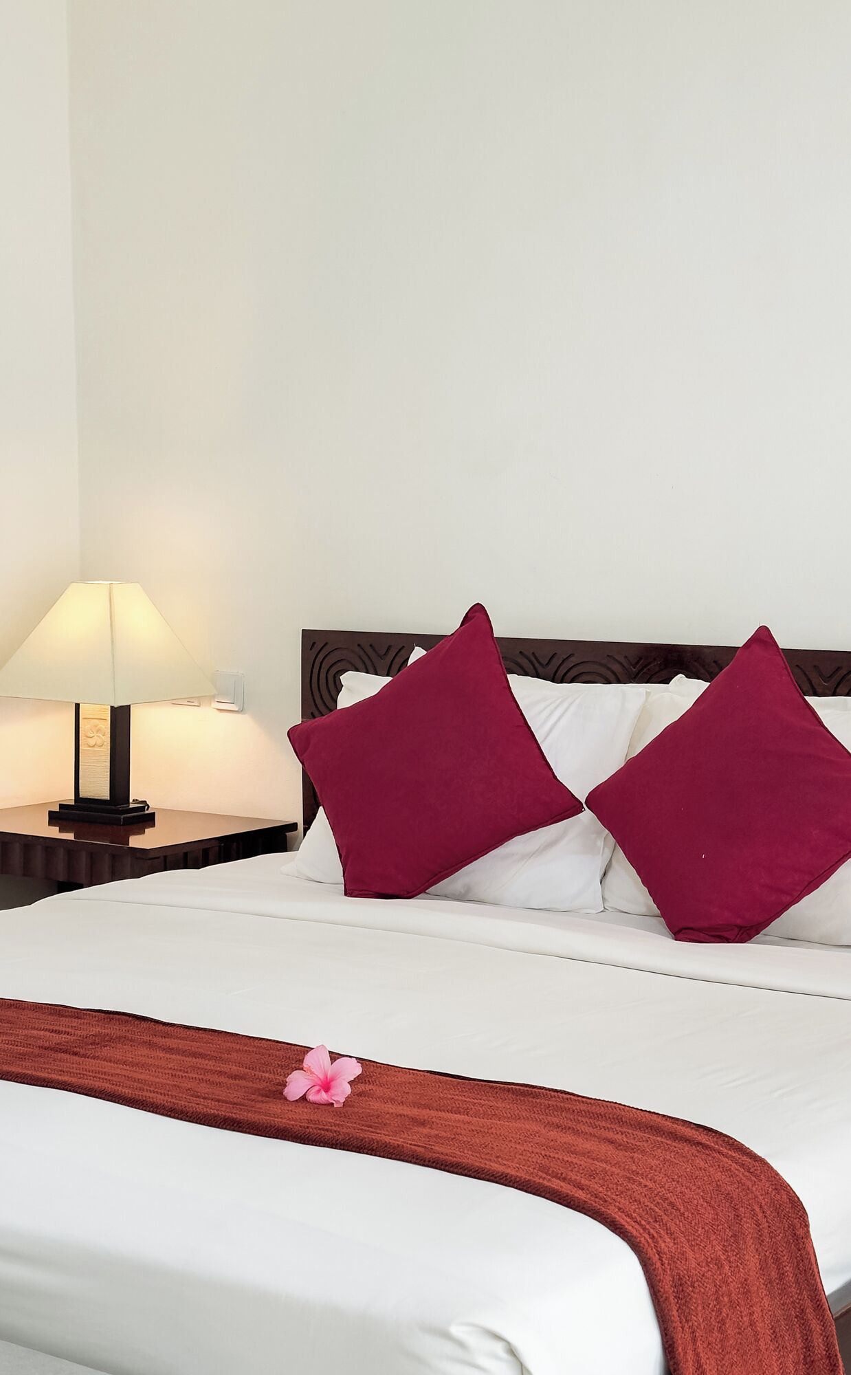 Una cama bien hecha con sábanas blancas y dos almohadas rojas, una lámpara de noche y una mesita de madera.