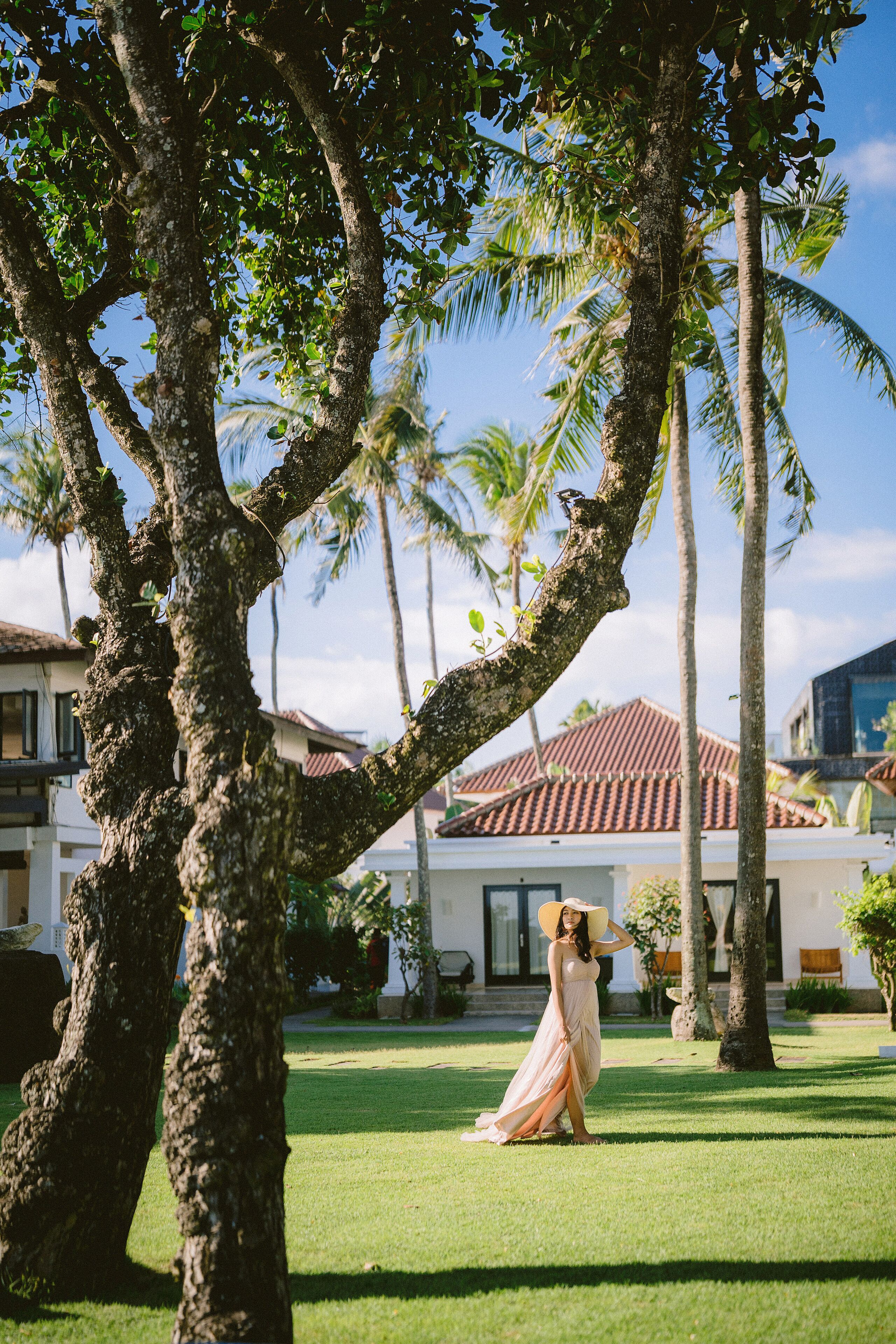 A woman in a long dress standing in a grassy area with palm trees and a building in the background.