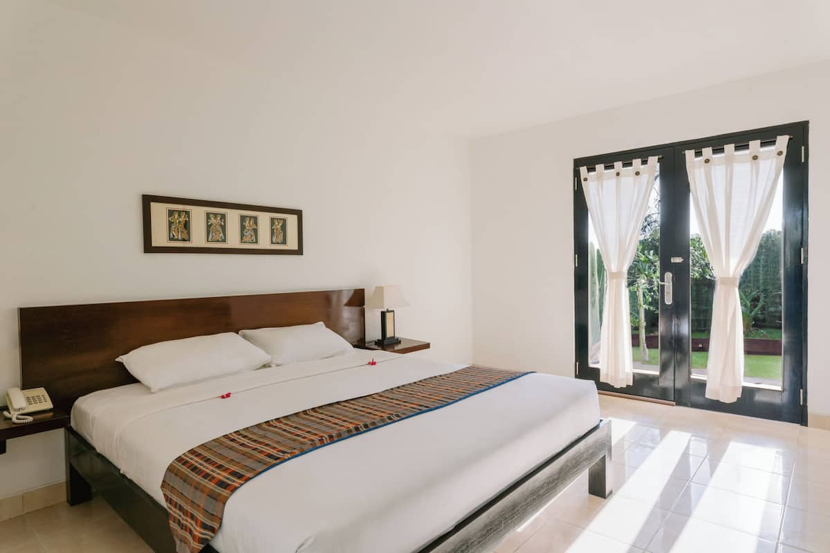 A hotel room with a large bed, a bedside telephone, a nightstand with a lamp, and a view through a glass door.