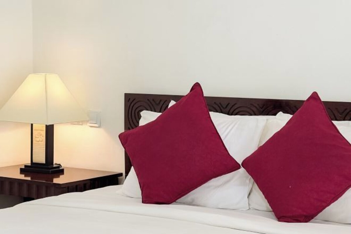 Una cama bien hecha con ropa de cama blanca y dos almohadas rojas, una lámpara de noche y un cabecero de madera.