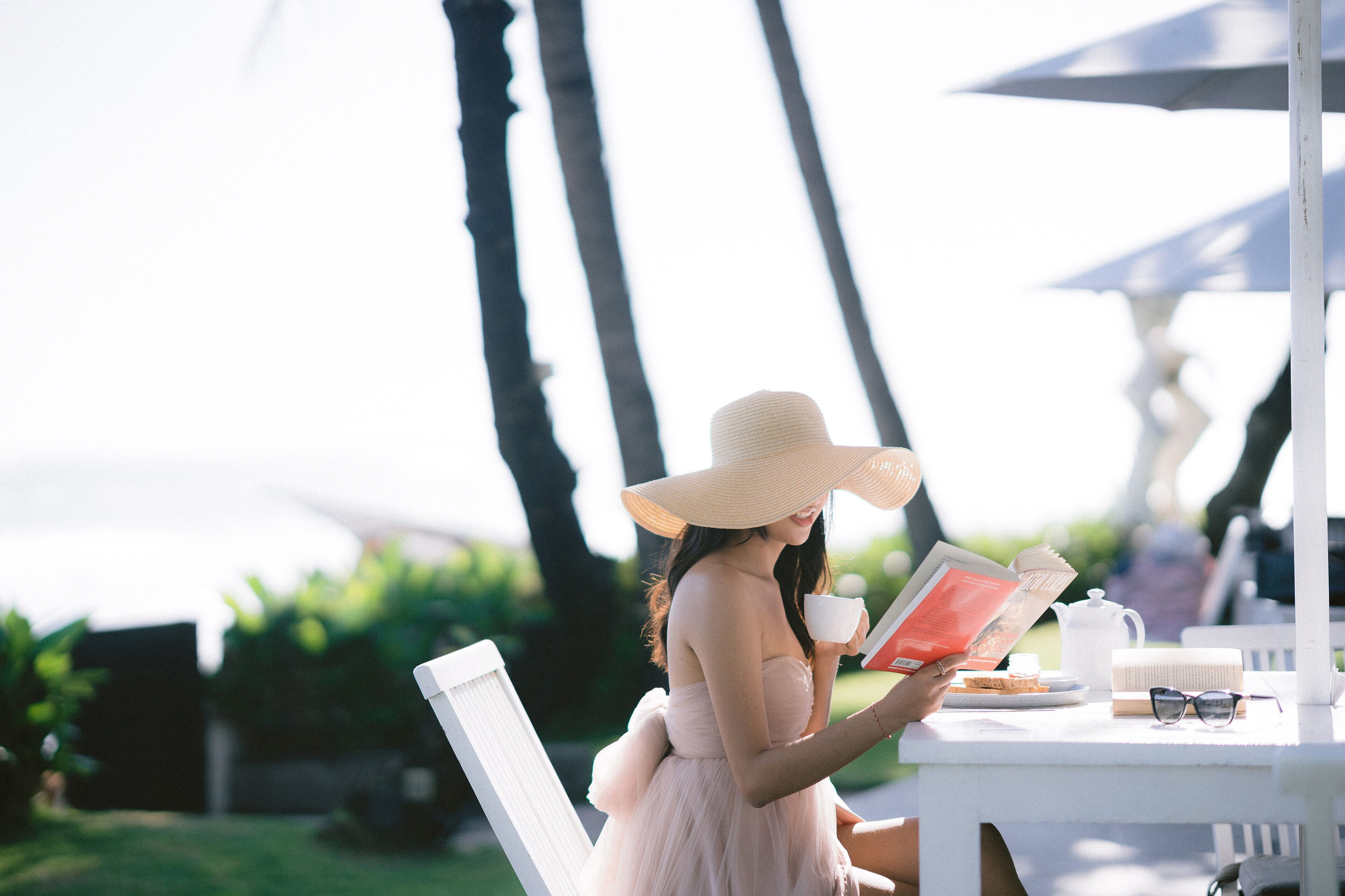 A person seated at a white table outdoors, reading a book, with a cup and a teapot on the table.