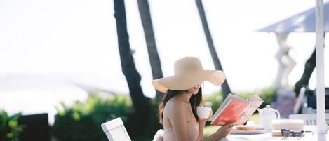 A person seated at a white table outdoors, reading a book, with a cup and a teapot on the table.