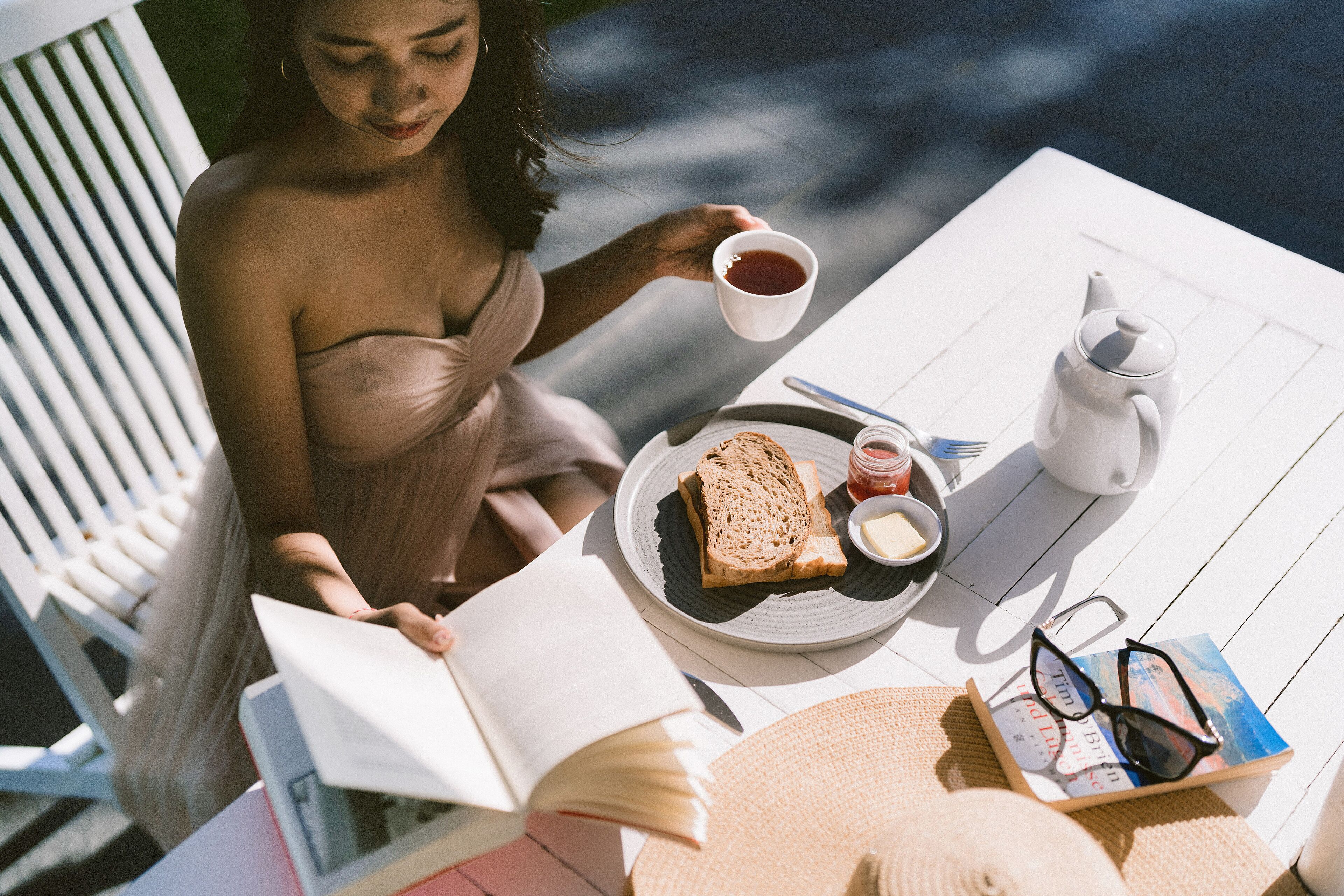 A person reading a book at a table with a plate of toast, butter, jam, and a teapot.