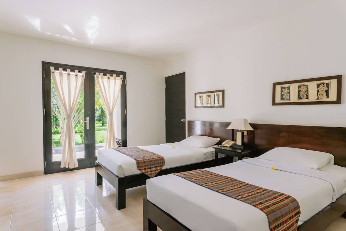 A hotel room with two beds, a door, a nightstand with a lamp, and framed pictures on the wall.