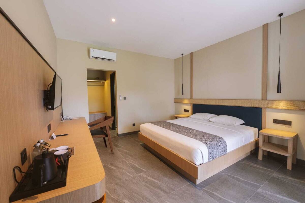 A hotel room with a bed, bedside tables, a desk with a chair, an air conditioning unit, and a television.