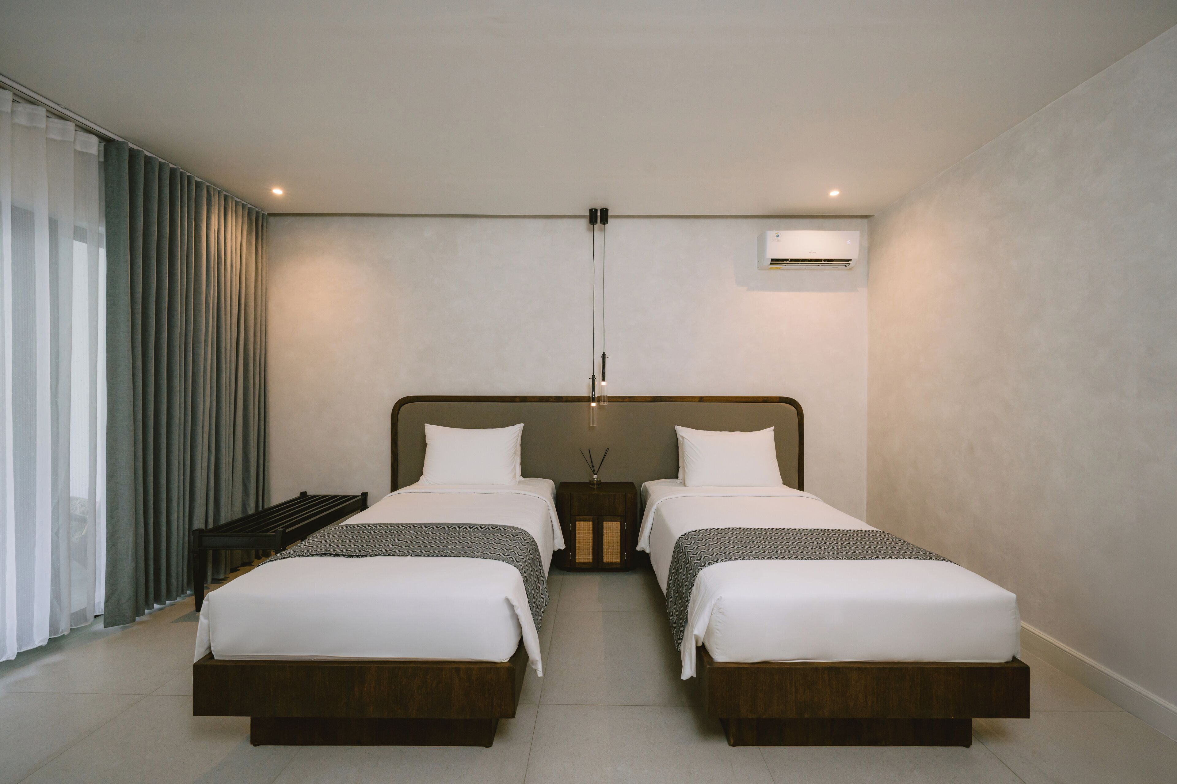 A modern hotel room with two single beds, a large window with curtains, a bedside table, and an air conditioning unit.