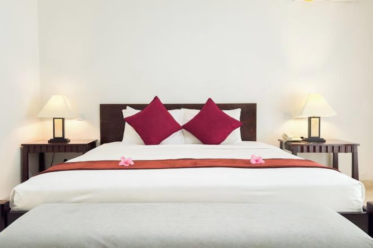 A neatly made bed with a wooden headboard, two red pillows, and a white bedspread. There are two bedside tables with lamps and a small clock on one of them.