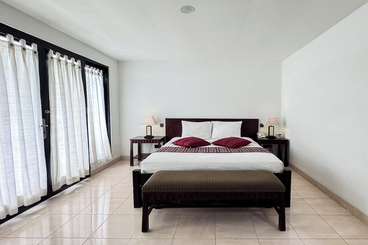 A modern hotel room with a large bed, two bedside tables with lamps, a bench at the foot of the bed, and a large window with curtains.