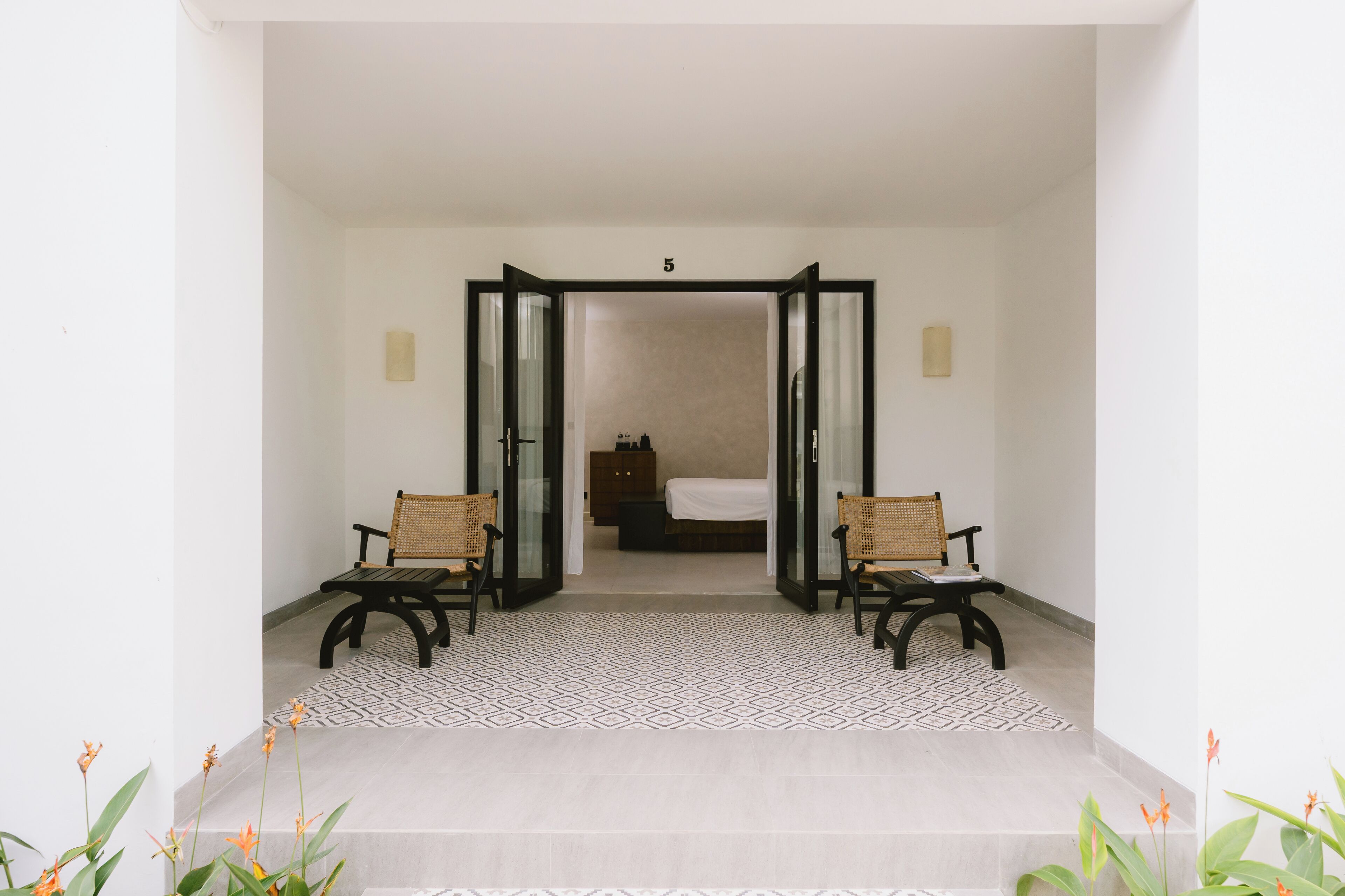 A hotel entrance with a modern design, featuring a geometric patterned floor, two wicker chairs, and a visible room number 5 in the background.