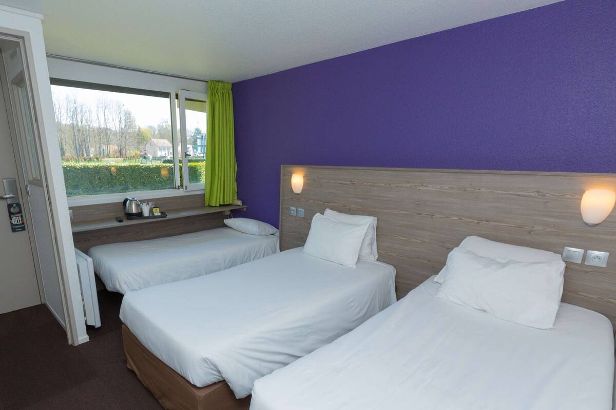 Quarto de hotel com duas camas de solteiro, janela com vista para vegetação, cabeceira de madeira e mesa de cabeceira com chaleira.