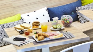 Desayuno con tostadas, cruasanes, jamón, queso, kiwi y un vaso de jugo de naranja sobre una mesa de madera.
