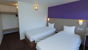Habitación de hotel con dos camas individuales, cabecera de madera, pared de acento morada y una pequeña estantería con luz.