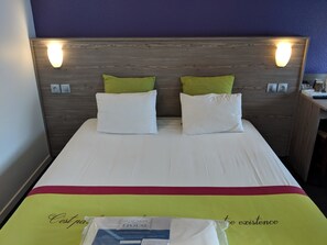 A hotel room with a wooden headboard, two white pillows, a green pillow, and a bedspread with a red and yellow border. - Kyriad Chantilly Sud - Luzarches (Chaumontel)