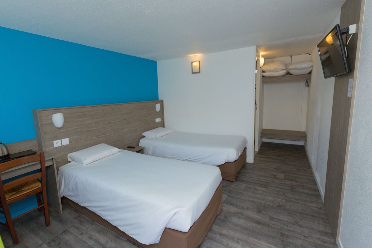 Quarto de hotel com duas camas de solteiro, uma escrivaninha com cadeira, televisão fixada na parede e uma pequena janela.