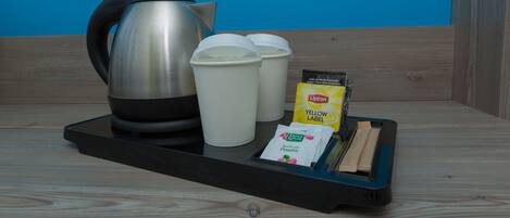Una bandeja con un hervidor de acero inoxidable, dos tazas blancas, un paquete de té Lipton amarillo y un estuche de perfume Saint Laurent sobre una superficie de madera frente a una pared azul.