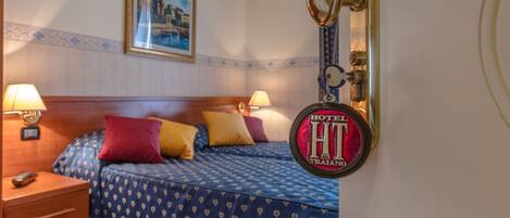 Una habitación de hotel con una cama, dos lámparas de noche, una mesita de noche, un cuadro en la pared y una puerta con cerradura y cartel.