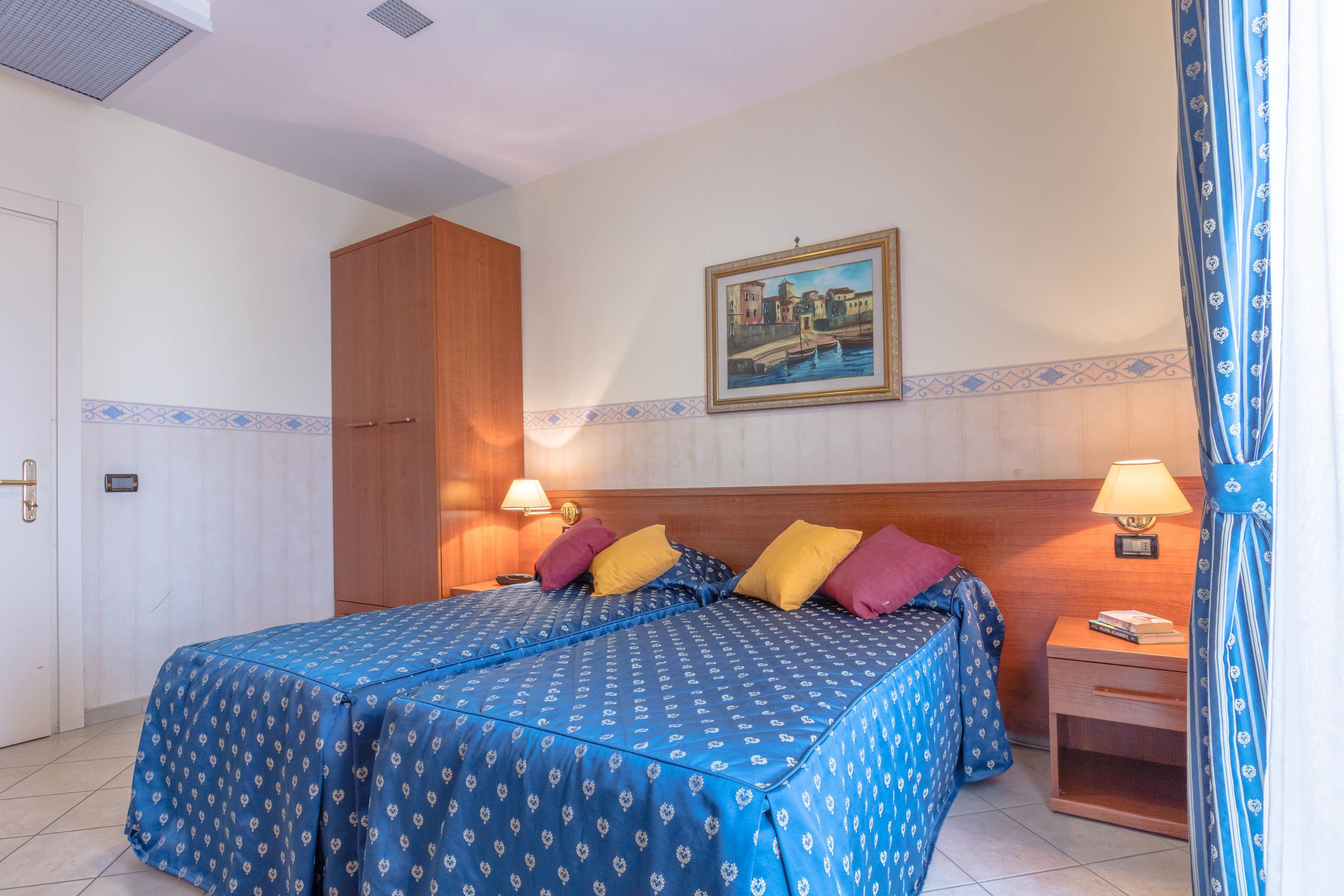 A hotel room with two beds, a wooden wardrobe, a nightstand with a lamp, and a painting on the wall.
