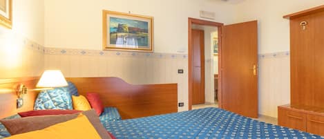 A hotel room with a bed, a wooden wardrobe, a painting on the wall, and a door leading to another room.