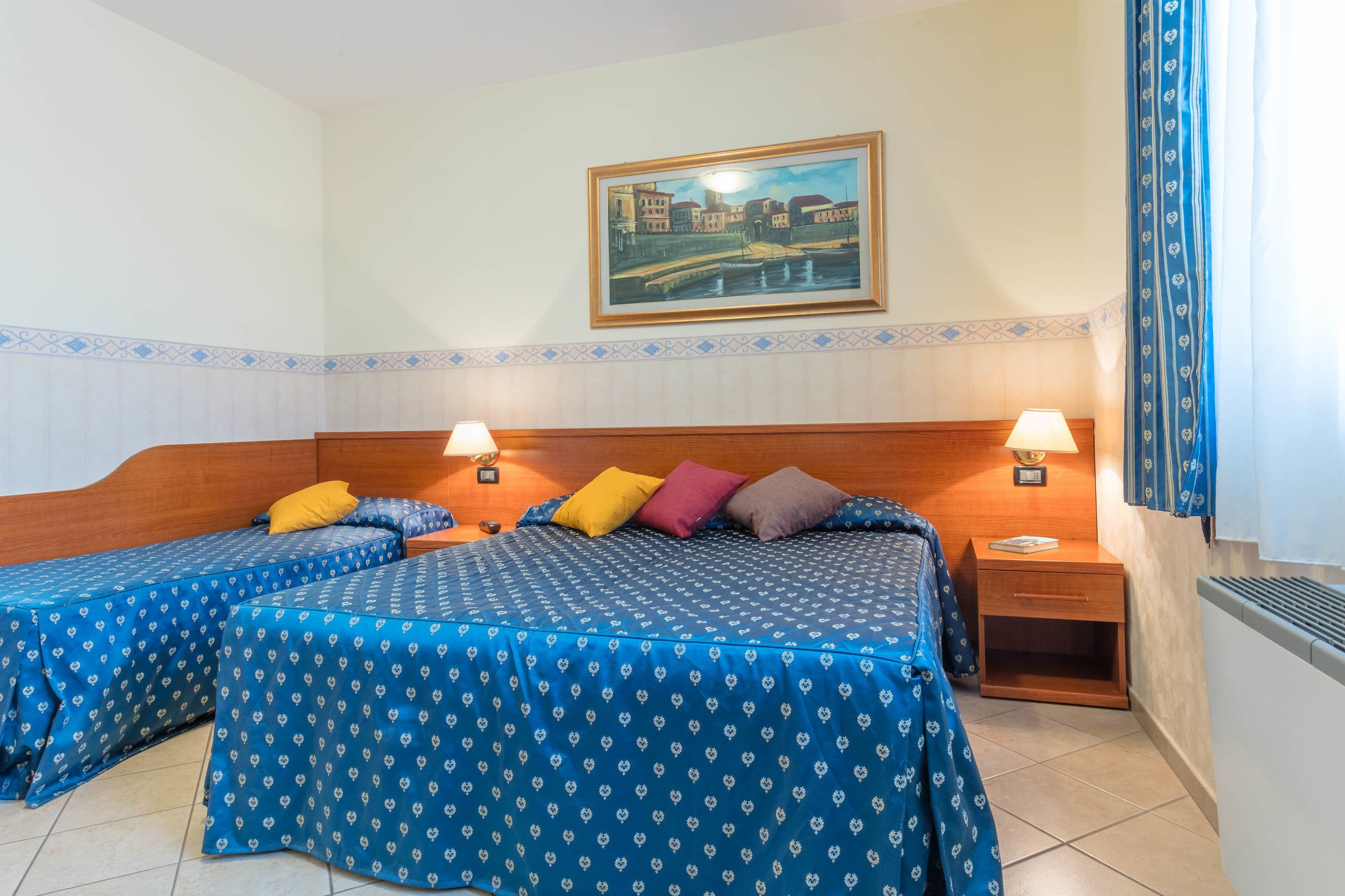 Ein Hotelzimmer mit zwei Betten, jeweils mit einer blau gemusterten Tagesdecke, einem Holzkopfstück und je zwei Nachttischlampen. Über den Betten hängt ein gerahmtes Gemälde einer Stadtansicht.